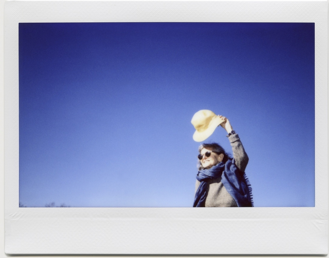 A woman stands outdoors and hold her hat to shield her from the sun